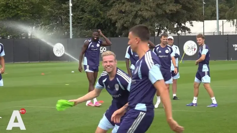 Voetballers van Ajax in blauwe trainingspakken trainen op een veld. Op de achtergrond sproeit een sproeier water terwijl de spelers rondlopen. Een speler op de voorgrond houdt een groen voorwerp vast, en een andere loopt langs hem heen. Het AT5-logo staat