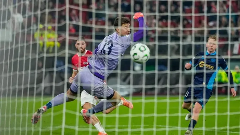 Een voetbalkeeper in een paars tenue duikt om de bal voor het doel te vangen, terwijl spelers van beide teams toekijken. Het tafereel wordt tijdens een wedstrijd door het doelnet heen bekeken.