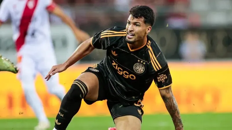 Een voetballer in een zwart Ajax-shirt met oranje biezen rent over het veld en lijkt te struikelen of te vallen. Op de achtergrond is een speler in een wit-rood tenue wazig te zien op een groen veld met een geel reclamebord.